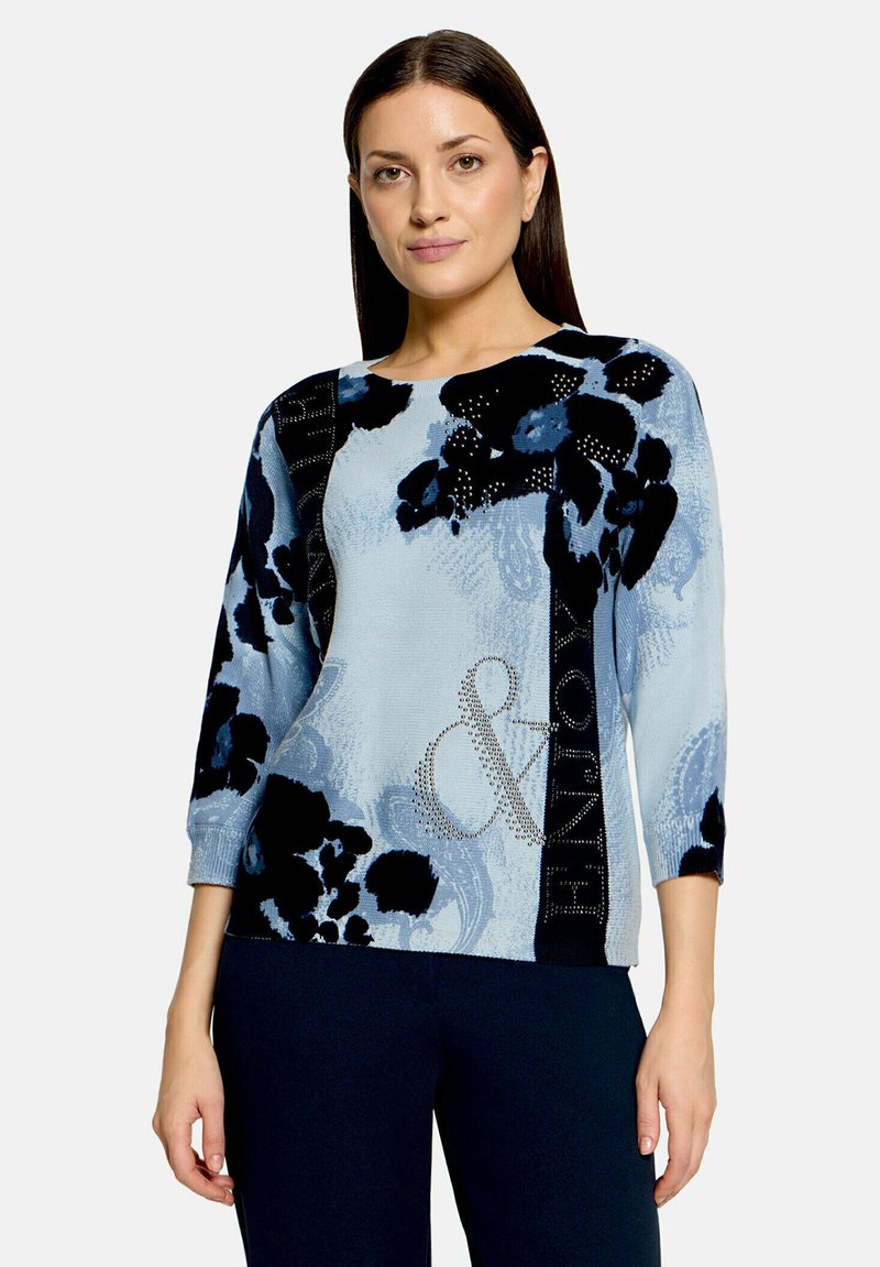 Femme aux cheveux lisses et foncés porte un pull bleu clair avec de grands motifs floraux noirs et une esperluette en strass, associé à un pantalon foncé.