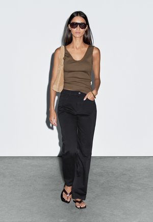 Braunes, tailliertes Tanktop, schwarze, weit geschnittene Hose und schwarze Riemchensandalen. Das Model trägt eine beigefarbene Schultertasche und oversize Sonnenbrillen.