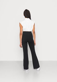 Camisa branca cropped com detalhe nos ombros, combinada com calças pretas de cintura alta e flair com riscas laterais. Modelo a usar sapatilhas brancas.
