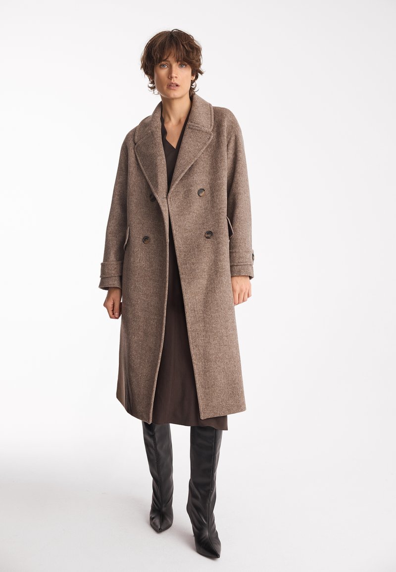 Brązowy płaszcz z wełny z podwójnym zapięciem, mający wycięty kołnierz, kieszenie nakładane i detale z guzikami, zestawiony z ciemnymi butami za kolano.