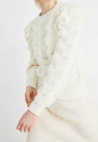 Pull blanc en tricot avec motif torsadé, manches bouffantes texturées et ourlet côtelé. Associé à une jupe assortie de couleur crème claire.