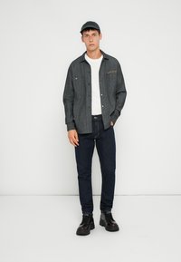 Camicia a righe scure sopra una maglietta bianca, abbinata a jeans blu scuro e scarpe nere, completata con un cappellino coordinato.