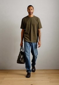 Bărbat care merge în interior, purtând un tricou verde măsliniu, blugi albaștri, pantofi maro, ținând o geantă de voiaj verde măsliniu în mâna stângă.
