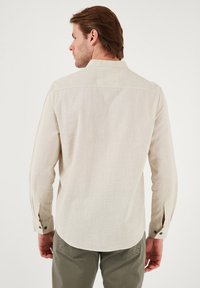 Chemise à manches longues beige clair avec un tissu texturé, un petit col et des poignets boutonnés. Le dos présente un style sans couture avec une légère courbure.