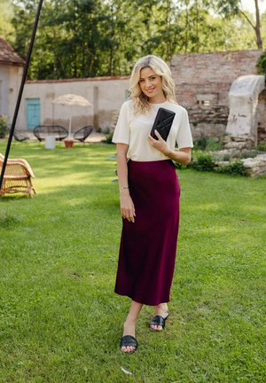 Žena v bílém krátkém rukávu a vínové midi sukni, která drží černou kabelku, stojí na zelené trávě v zahradním prostředí.