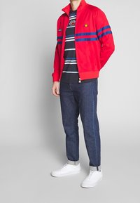 Chaqueta roja con cremallera y rayas horizontales azules, usada sobre una camisa de rayas oscuras. Combinada con jeans oscuros y zapatillas blancas.