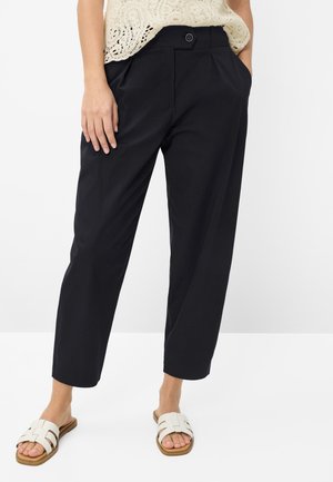 Vrouw draagt zwarte cropped broek met knoop aan de voorkant en zakken, crèmekleurige kanten top en witte slippers, staand met hand in de zak.