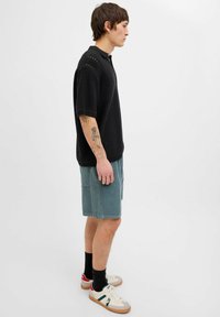 Tricou polo din tricot negru cu mâneci scurte, asortat cu pantaloni scurți din catifea de culoare teal. Încălțămintea include adidași albi cu accente verzi și roșii.