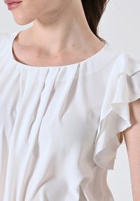 Primo piano di una donna che indossa una blusa senza maniche bianca con dettagli arricciati sulle spalle e pieghe sul collo.