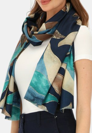 Kobieta ubrana w biały top i niebiesko-turkusowo-beżowy szal z metalicznymi akcentami, owinięty wokół szyi i ramion.