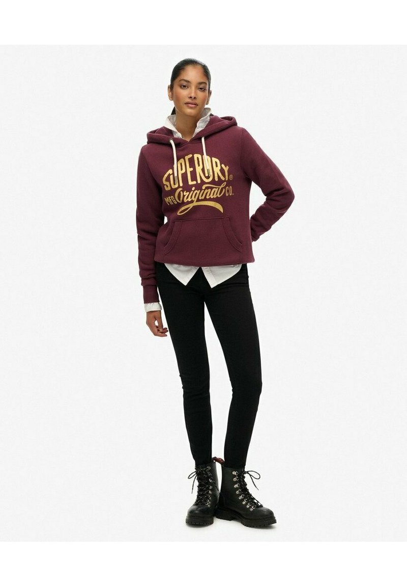 Bordowy krótki sweatshirt z złotym nadrukiem, z kapturem, sznurkami i kieszenią z przodu. W połączeniu z czarnymi obcisłymi dżinsami i butami na sznurówki.