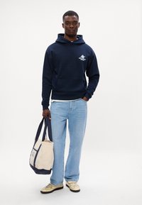 Hanorac bleumarin cu buzunar frontal și logo, blugi deschiși, geantă bej și bleumarin, și adidași bej cu accente închise.