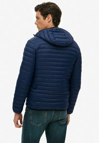 Mężczyzna w granatowej pikowanej kurtce z kapturem i niebieskich dżinsach, widziany z tyłu na jednolitym tle.