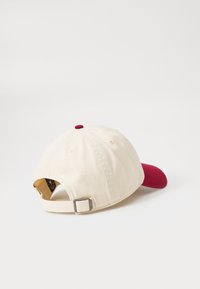 Cappellino da baseball in cotone con corpo crema e visiera rossa. Presenta una cintura in tessuto e una fibbia in metallo sul retro. Texture liscia, design semplice.