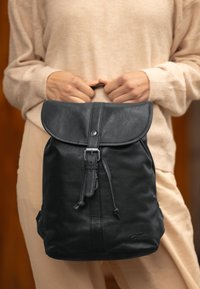 Mochila de cuero negro con una solapa redondeada, cierre de cordón y acentos metálicos. Textura suave con sutiles detalles de costura.