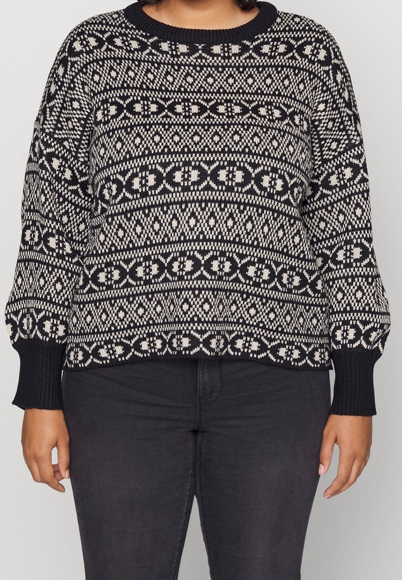 Pull à manches longues noir et blanc à motifs géométriques, porté avec un pantalon foncé, visible des épaules aux hanches.