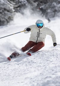 Skiër in een witte insulated jas en een bruine broek, carve in poedersneeuw. Hij draagt een helm en een bril met blauwe glazen.