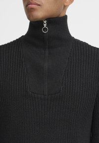Schwarzer gestrickter Pullover mit hohem Kragen und einem Halbreißverschluss auf der Vorderseite, der ein strukturiertes Muster und eine dezente, geformte Ausschnittlinie aufweist.