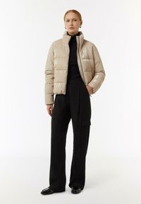Beżowa błyszcząca kurtka puchowa z wysokim kołnierzem, zapięciem na zamek i pikowanym wzorem, noszona na czarnym golfie i pasujących szerokich spodniach.