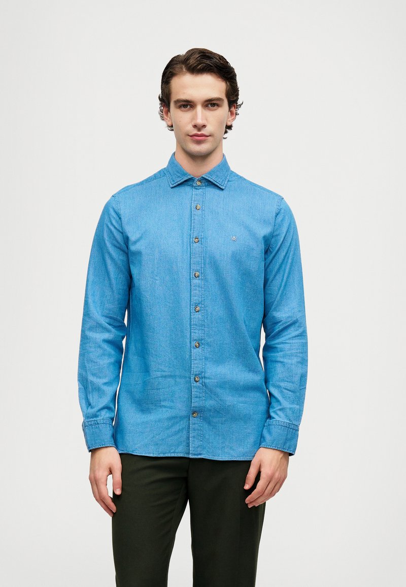 Camicia azzurra chiara a maniche lunghe con un tessuto testurizzato. Presenta un colletto a punta e sei bottoni frontali, abbinata a pantaloni scuri.