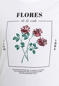 Bílý tričko s grafikou tří červených flitrových růží se zelenými listy, umístěných v černém obdélníkovém rámečku, spolu s textovými prvky.