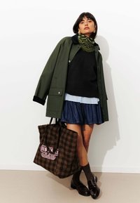 Manteau vert olive, pull noir, chemise bleu clair, jupe en denim, sac à carreaux marron avec texte rose, et chaussettes noires avec des chaussures.