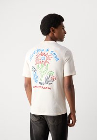 Krämfärgad t-shirt med korta ärmar som har en färgglad blommönster på ryggen, med blå, röda och gröna accenter; mjuk bomullstyg.