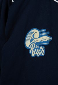 Tissu bleu avec un patch cousu représentant un casque ailé et le texte « in a Rush » en blanc et bleu.