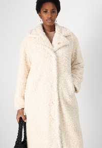 Crème-kleurige, fluffy jas met een sjawlboord, knoopsluiting en twee zijzakken. Model houdt een gestructureerde zwarte tas vast.