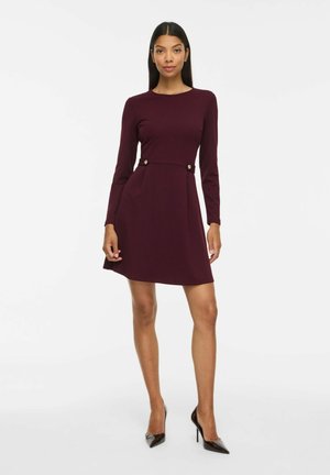Femme debout portant une robe à manches longues violet foncé avec des boutons à la taille et des chaussures à talons noirs sur fond blanc.