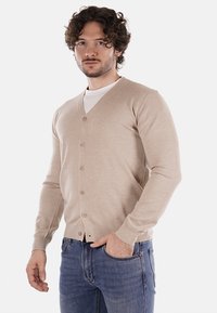 Cardigan beige chiaro con scollo a V, maniche lunghe e sei bottoni, realizzato in materiale morbido. Indossato sopra una camicia bianca e abbinato a jeans blu.