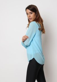 Blusa de manga larga azul claro con una textura translúcida y un dobladillo redondeado, combinada con leggings negros. Presenta un corte holgado y puños con botones.