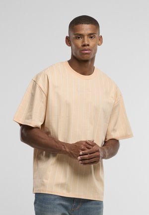 Jeune homme debout, les mains jointes, portant un t-shirt beige ample avec des rayures verticales blanches et un jean bleu sur un fond uni.