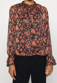 Sort bluse med orange blomsterprint, sammensamlet halslinje, lange ærmer med flæsemanchetter og let, gennemsigtig stof.