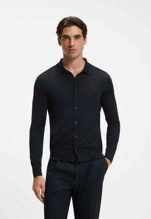 Hombre que lleva una camisa negra ajustada de manga larga con botones y cuello, y pantalones oscuros, de pie frente a un fondo claro y liso.