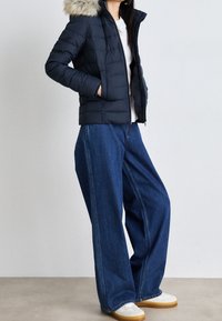 Navy puffer jacket with a fur-trimmed hood, paired with wide-legged denim jeans. Model wears white sneakers. Textured materials visible.