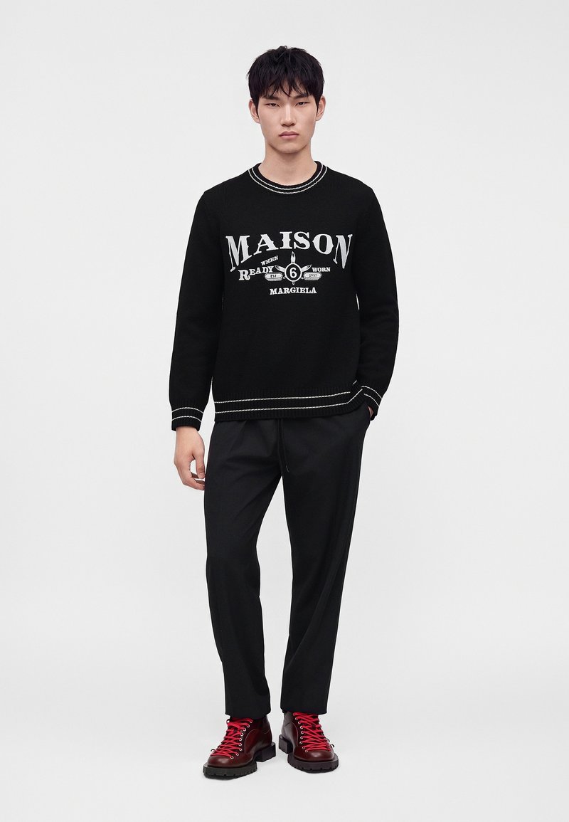Giovane uomo che indossa un maglione nero con testo bianco "Maison Margiela", pantaloni neri e stivali rosso scuro con lacci rossi in piedi su sfondo bianco.