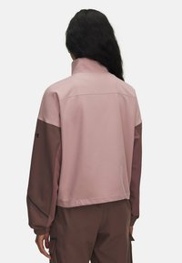 Person mit langen dunklen Haaren, die eine zweifarbige Jacke in Pink und Mauve sowie passende Hosen trägt, von hinten vor einem schlichten Hintergrund gezeigt.