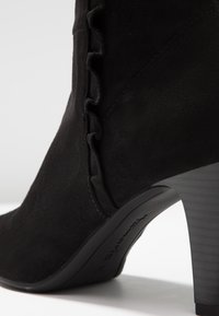 Schwarze Wildleder-Ankle-Boots mit einer Rüsche an der Seite, einem hohen Absatz und einer glatten Textur an Obermaterial und Sohle.