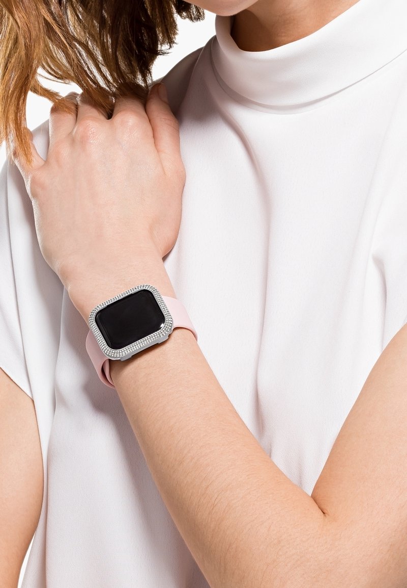 Femme portant un haut blanc avec une montre connectée rose à écran noir et un cadran orné de strass sur son poignet gauche.