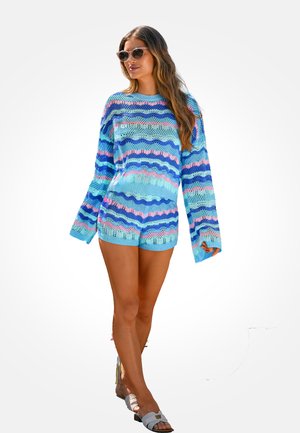 Vrouw draagt een blauw, roze en paars gestreepte gebreide trui en bijpassende korte broek, zonnebril en witte sandalen, staand tegen een witte achtergrond.