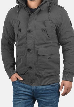 Homme portant une veste à capuche gris foncé avec des poches boutonnées et un jean bleu, une main dans la poche et l’autre le long du corps.