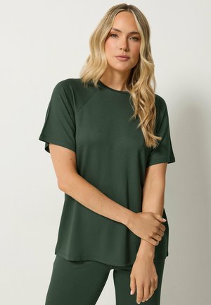Femme blonde portant un haut à manches courtes vert foncé et un pantalon assorti, debout les bras légèrement croisés devant un fond clair uni.