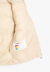 Indersiden af en beige dunjakke med quiltet tekstur. Indeholder en lomme med tags, der viser farverige mærkater og plejeinstruktioner. Lynlås synlig.