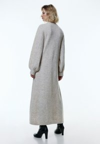 Langes graues Strickkleid mit langen Ärmeln und lässigem Schnitt, ausgestattet mit einem runden Ausschnitt und seitlichen Schlitzen, getragen mit schwarzen Stiefeletten mit Absatz.