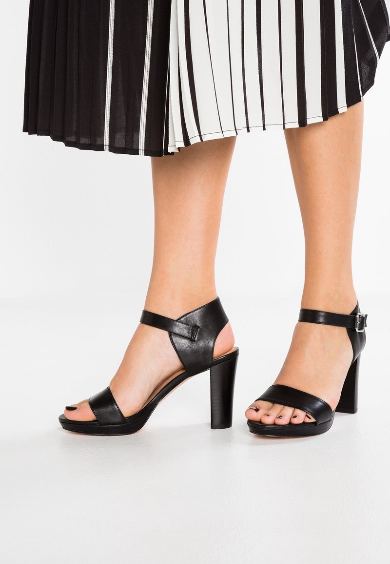 Jambes portant une jupe plissée noir et blanc avec des sandales à talons hauts en cuir noir, avec des brides à la cheville et des orteils découverts.