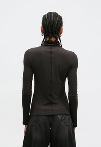 Zwart nauwsluitend shirt met lange mouwen van zachte stof, met een hoge hals en een centrale naad. Draagt losse, donkere spijkerbroek.