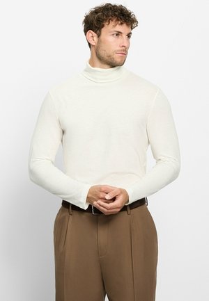 Mann mit lockigem Haar, der einen weißen Rollkragenpullover und braune Hose trägt, steht mit verschränkten Händen da und schaut zur Seite.