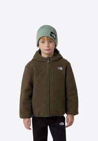 Pruun fliisist lukuga jakk kapuutsiga, mida täiendab mintroheline soonikuga müts. Mustad püksid valge logoga jalal.