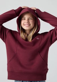 Fată tânără zâmbind cu mâinile pe cap, purtând un hanorac maroniu Levi's și blugi blue, pe un fundal simplu deschis.